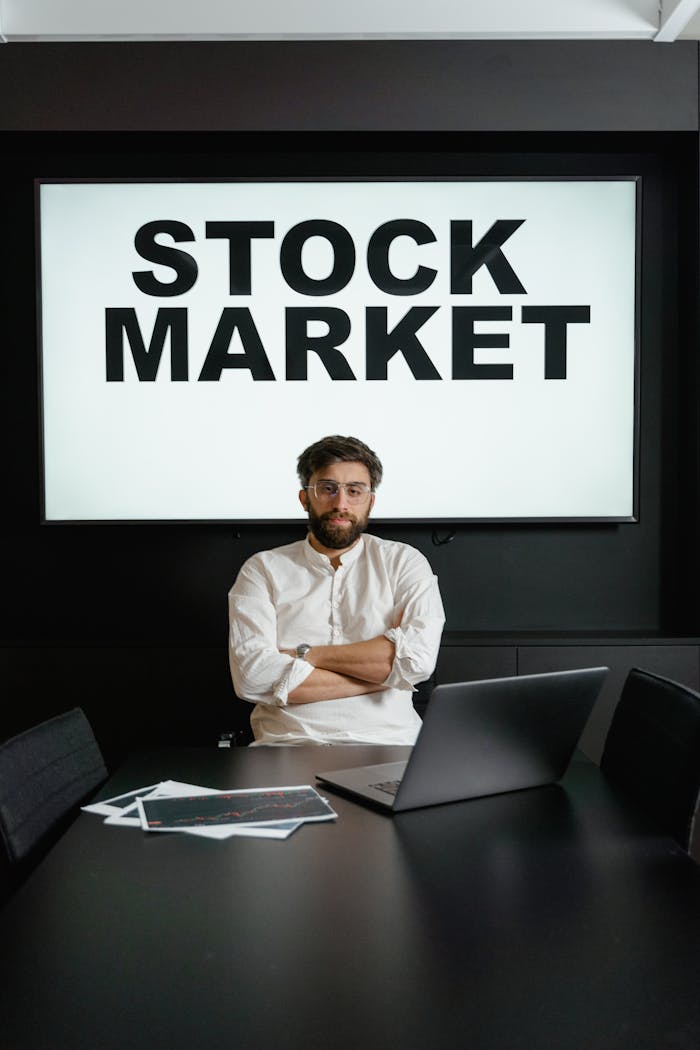 Professional businessman with arms crossed in front of a stock market display in a modern office setting.